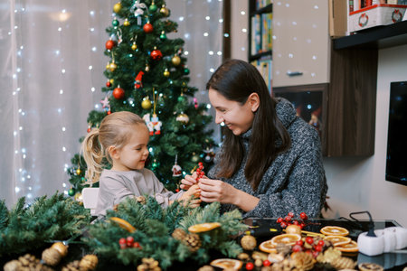 Smiling mother showing little girl berries for Christmas wreath while sitting at tableの写真素材
