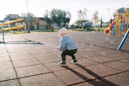 Little girl squats down on the playground and catches bubbles. Back viewの写真素材