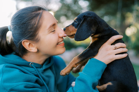Black puppy licks the nose of a smiling girl holding him in her armsの写真素材