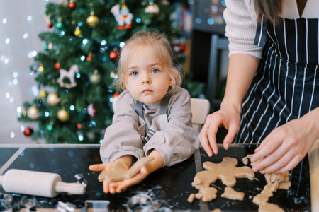 Little girl holds a cut out cookie on her outstretched palm while sitting next to her mother sorting raw cookies on the tableの写真素材