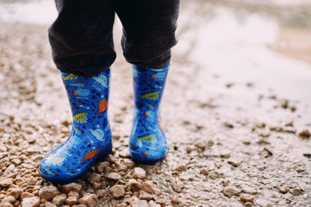 Small child in colorful rubber boots stands on gravel in a puddle. Cropped. Facelessの写真素材