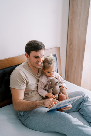 Smiling dad reading a book to a little girl sitting with a teddy bear on his lap on the bedの写真素材