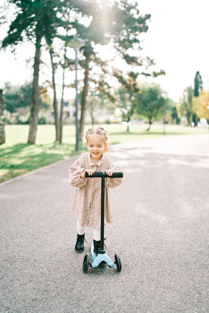 Little smiling girl rides a scooter, pushing off with her foot on the road in the parkの写真素材