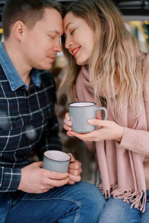 Smiling man and woman touching foreheads while sitting with cups in their handsの写真素材