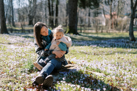 Mom looks at a little girl on her knees sitting on a stump in a clearing with blooming flowersの写真素材
