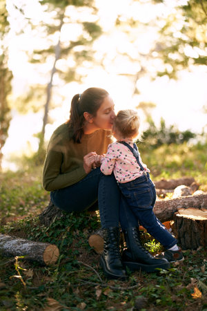 Mom sits on a stump and kisses a little girl standing next to her in the forestの写真素材