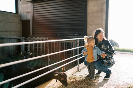 Smiling mother squatting near little girl at farm next to goat paddockの写真素材
