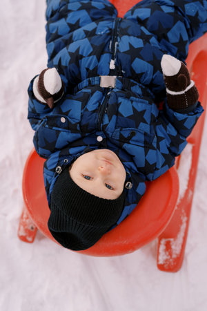 Little boy in a jacket and hat lies on a sleigh. Top viewの写真素材