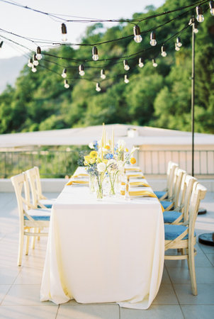 Long festive table with bouquets of flowers on the terrace with hanging garlands of light bulbs