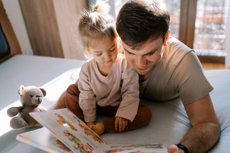 Smiling dad reads a book to a little girl sitting on the bed. Top viewの写真素材