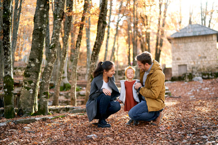 Dad and mom are squatting and looking at a little squinting girl in the autumn forestの写真素材