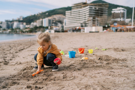 Little girl on the beach pours sand with a toy shovel into a plastic bucket while squattingの写真素材