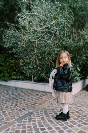 Little girl with a toy hare stands on a path near an olive tree and looks to the side, touching her chinの写真素材