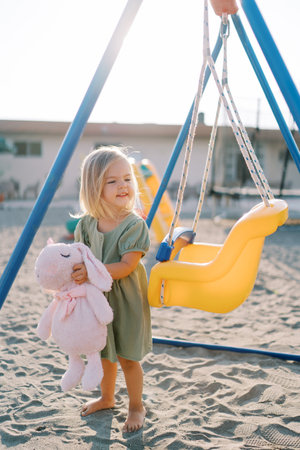 Little smiling girl stands hugging a pink rabbit near a swing on the playground and looks awayの写真素材