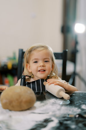 Little smiling girl sits at a table with a rolling pin near the doughの写真素材
