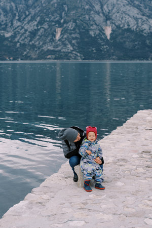 Mom squatted next to a little girl sitting on a bollard on the pier