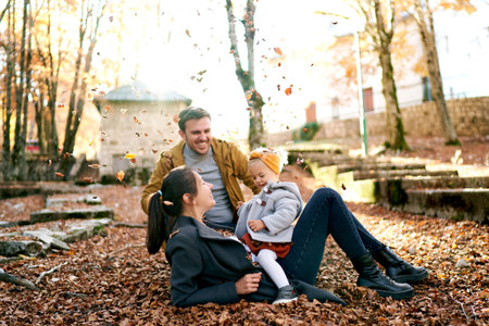 Dad sits next to mom lying on the ground with a little girl on her stomach under falling leaves