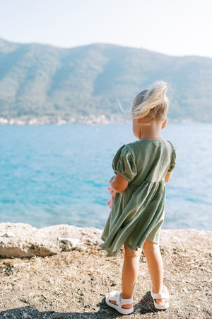 Little girl with a soft toy stands in a dress fluttering in the wind on a rock above the sea. Back viewの写真素材