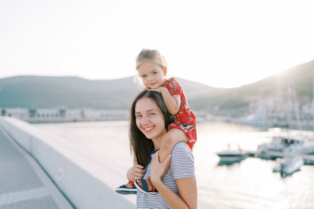 Little girl sits on the shoulders of a smiling mother, resting her chin on her head on the seashoreの写真素材