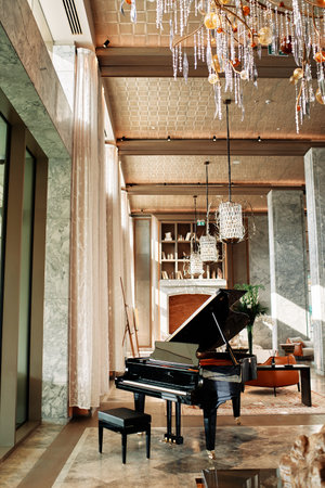 Black piano with a stool stands in a hotel lobby with modern chandeliers hanging from the ceilingの写真素材