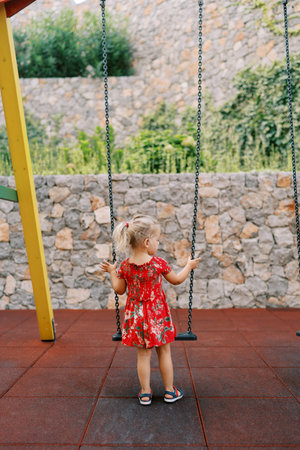 Little girl stands by a chain swing on a playground near a stone wall covered with ivyの写真素材