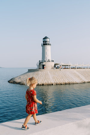 Little girl walks along the embankment overlooking the lighthouse on the breakwater. Back viewの写真素材