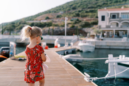 Little girl with a pink toy walks along the pier past the moored yachts. Back view