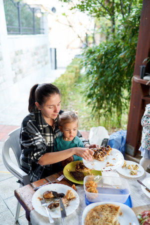 Mom takes a treat from the plate at the table while sitting with a little girl in her arms on a chairの写真素材