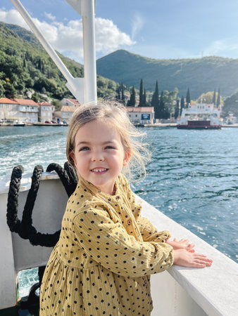 Little girl stands leaning on the side of a boat floating on the sea towards a mountainous shoreの写真素材