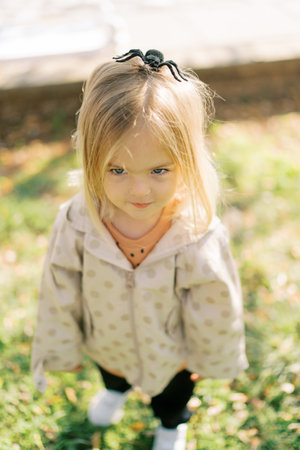 Little girl with a toy spider on her head stands on a green flowerbed