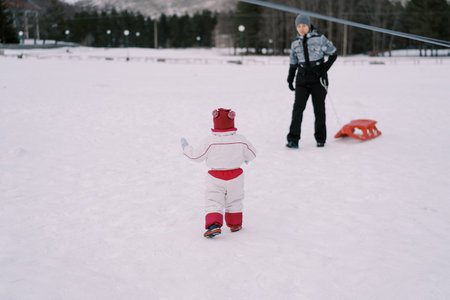 Little girl goes to her mother standing with a sled in the snow. Back viewの写真素材