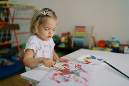Little girl draws in an album with a brush with paints sitting at the tableの写真素材