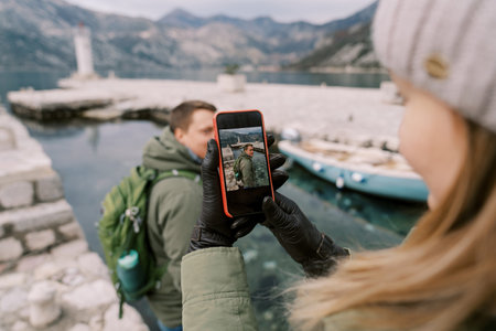 Girl takes a picture on a smartphone of guy on a pier by the sea. Side viewの写真素材