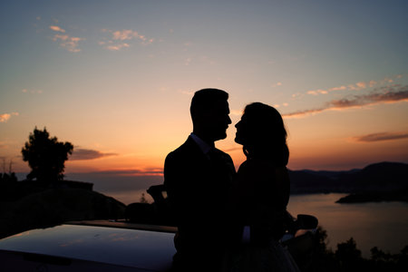 Bride and groom are hugging while looking at each other near a car in the mountains at sunsetの写真素材