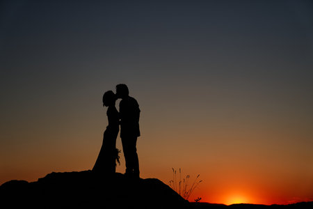 Silhouettes of bride and groom kissing while standing on the mountain at sunsetの写真素材