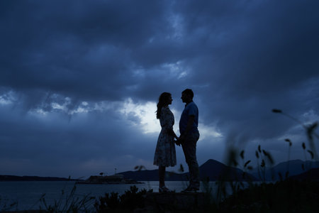 Man and woman stand holding hands by the sea against a dark skyの写真素材