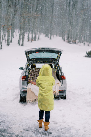 Woman stands near a car with an open trunk and a spread blanket inside in a winter park. Back viewの写真素材