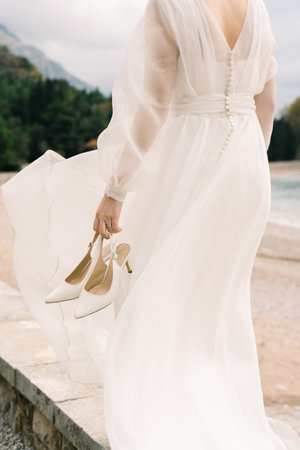 Bride with shoes in her hand walks along the fence on the seashore. Back view. Croppedの写真素材