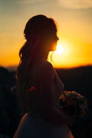 Bride with a bouquet stands in the rays of the setting sun in the mountains. Side viewの写真素材