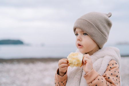 Little girl with an apple in her hands stands by the sea and looks into the distanceの写真素材