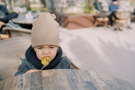 Little girl eats porridge with a spoon while sitting at a table on a chair in a street cafeの写真素材