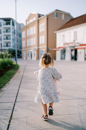 Little girl with a pink toy rabbit walks along the road to an apartment building. Back viewの写真素材