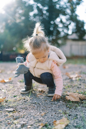Little girl with a soft toy squats down on a gravel path and touches a pebble with her fingerの写真素材