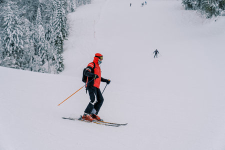 Skier in a red ski suit turns from a side track onto a ski slope along a snowy forest