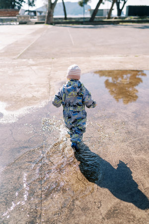 Little girl in overalls walks through a puddle, making splashes. Back viewの写真素材