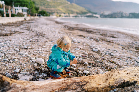 Little girl squats and plays with pebbles near driftwood on the beachの写真素材