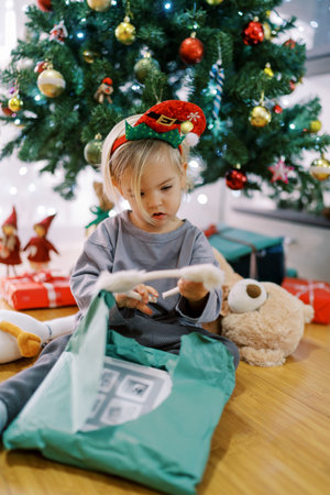 Little girl in a hoop with a gnome hat examines a hoop with cat ears, sitting by the Christmas treeの写真素材
