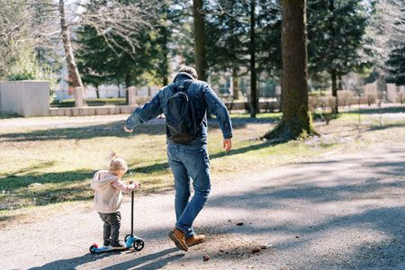Dad is carrying a little girl on a scooter along a park road on a rope. Back viewの写真素材