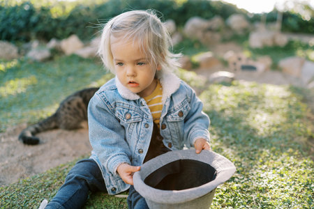Little girl with a hat in her hands sits on green grass and looks awayの写真素材