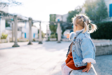 Little girl sits on a stone fence in the yard and points her finger into the distance. Side viewの写真素材